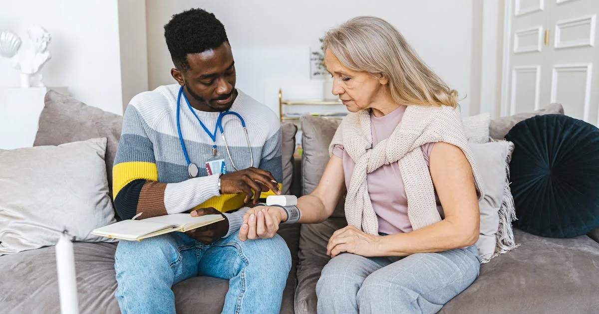 A male healthcare worker with stethoscope checks a senior woman's pulse oximeter while seated together on a couch in her home living room